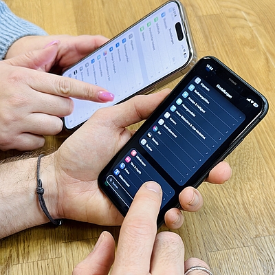 Die Hände zweier Personen halten jeweils ein iPhone in der Hand und bedienen die Einstellungen. Auf einem Gerät sind die iOS-Einstellungen im hellen Modus zu sehen, auf dem anderen im dunklen Modus.