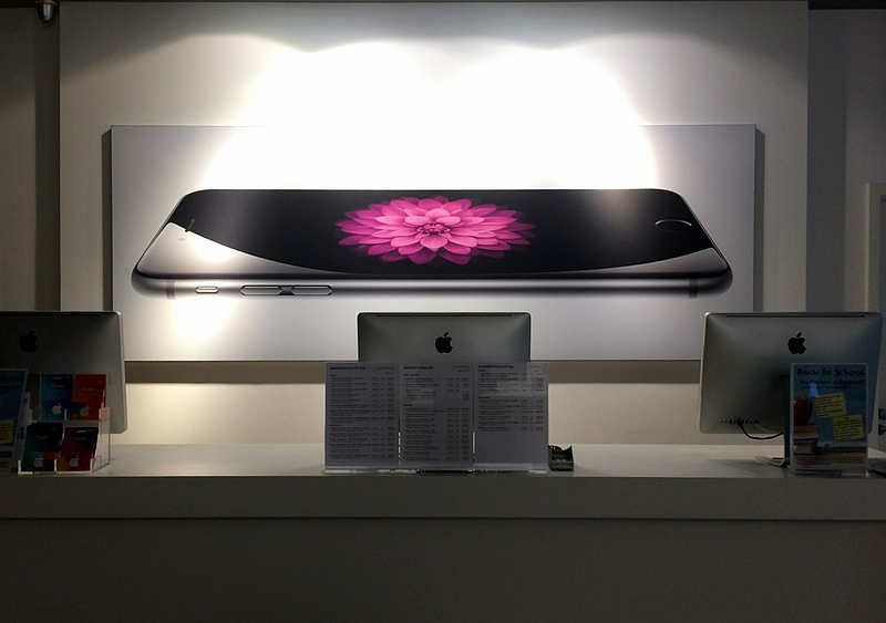 hinter einer Theke auf der drei Monitore von hinten zu sehen sind, ist ein Werbeposter mit einem großen horizontal liegendem iPhone 6 