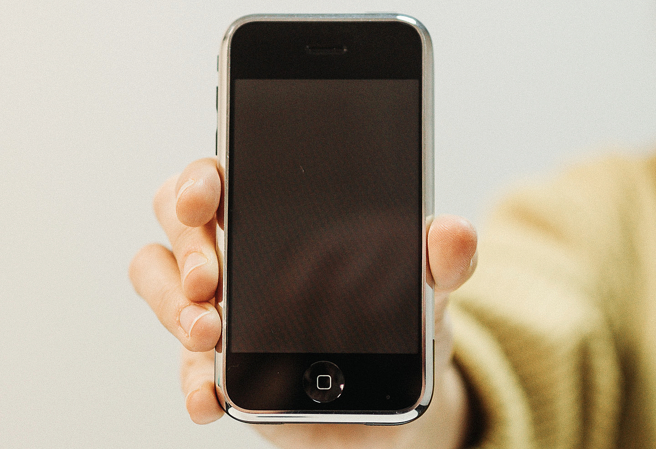 eine Hand hält einem das iPhone der ersten Generation mit dem Bildschirm entgegen