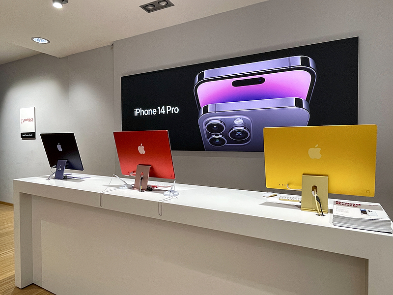 Theke mit drei iMacs, dahinter Werbeposter mit iPhone 14 Pro