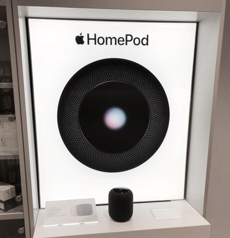 ein schwarzer HomePod steht vor einem beleuchteten Werbeposter auf dem die kreisrunde HomePod Oberfläche zu sehen ist